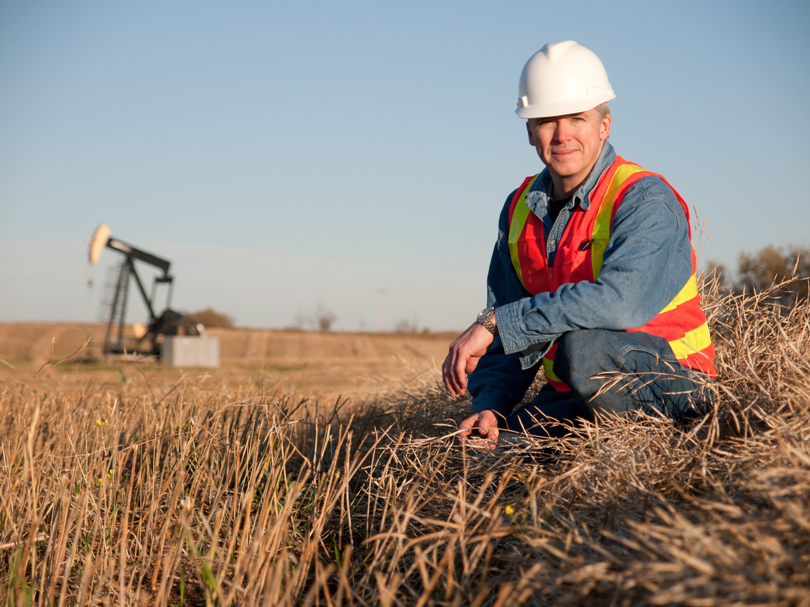 a man kneeling in a field with an oil pump in the background