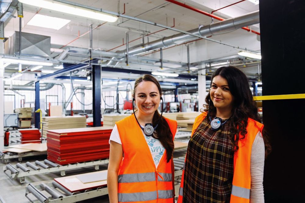 two women wearing orange vests are standing in a factory