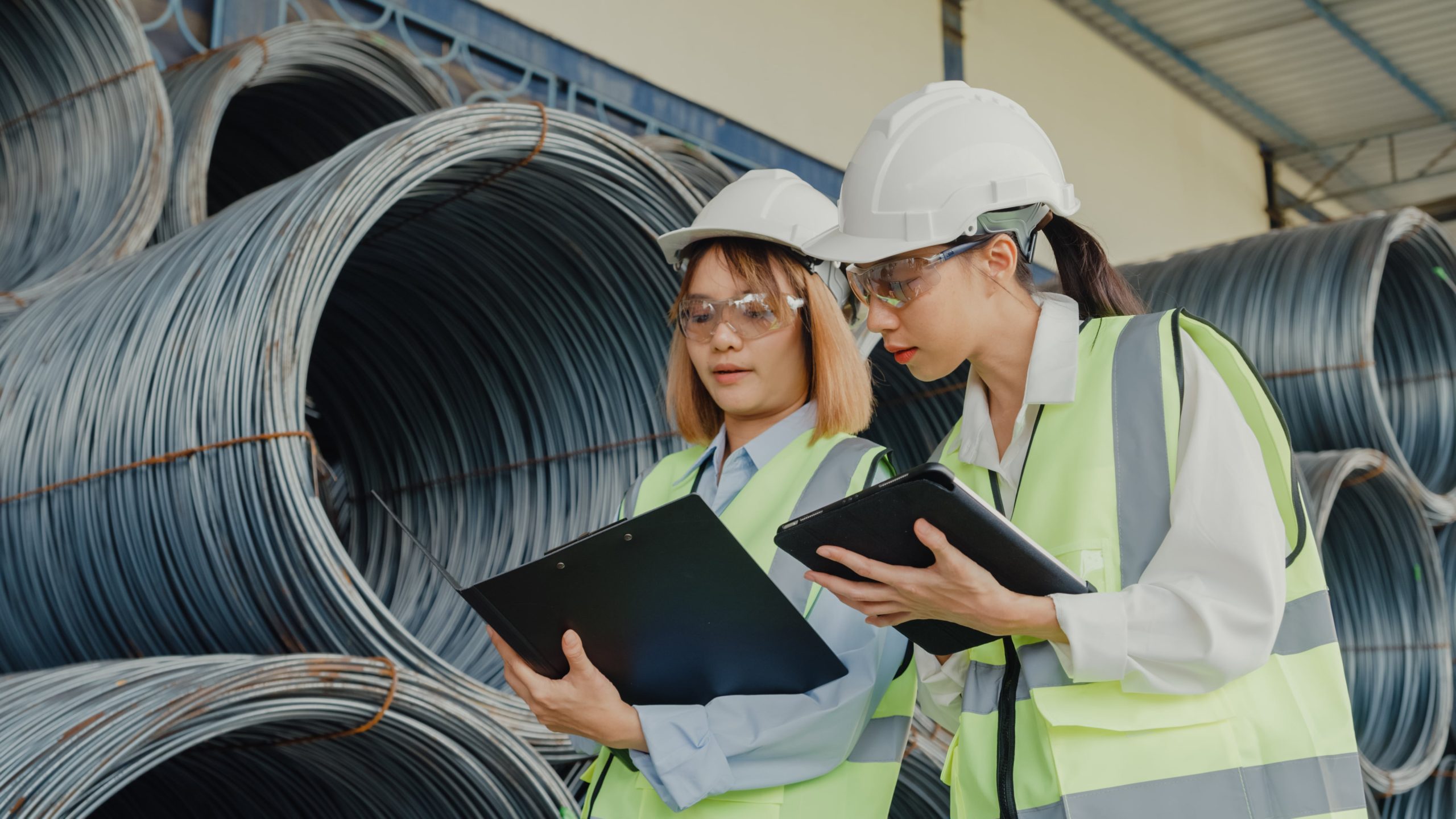 two women wearing hard hats and safety vests look at a clipboard